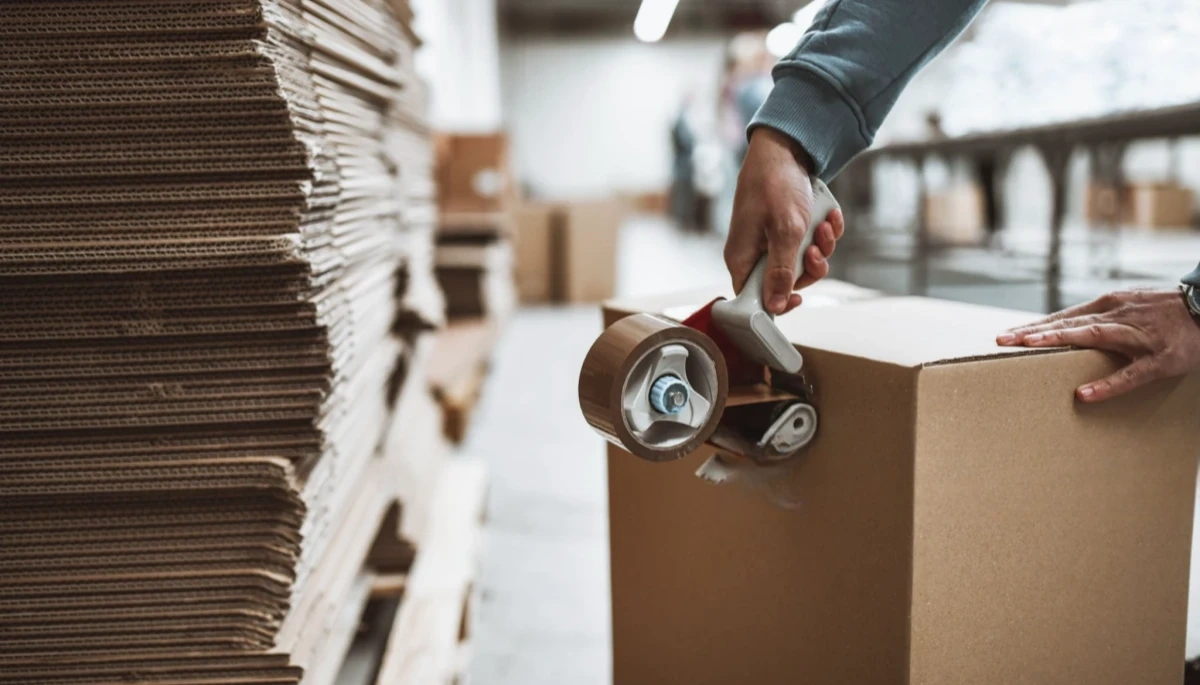 Worker sealing a cardboard box with tape in a warehouse, demonstrating our commitment to fast and secure packaging and delivery services at Smart Innovation.